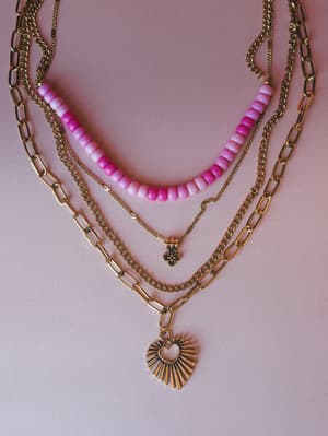 A layered necklace set styled against a dusty pink background, mixing gold chain links, a pink beaded strand with ombre detail, a delicate cross charm chain, and a bold paperclip chain with a sunburst heart pendant.