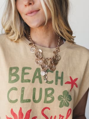 A close-up of a gold coin charm necklace styled over a sandy tan 'Beach Club' graphic tee, the layered medallion chains feeling effortlessly summery and a little collected — like something you'd find at a seaside market and never take off.