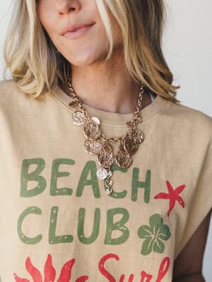 A close-up of a gold coin charm necklace styled over a sandy tan 'Beach Club' graphic tee, the layered medallion chains feeling effortlessly summery and a little collected — like something you'd find at a seaside market and never take off.