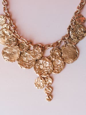 A close-up of a hammered gold coin charm necklace layered on a soft blush background — the kind of piece that looks like it traveled home from a market in Greece.
