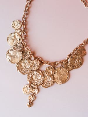 A statement gold coin necklace with hammered, textured disc charms clustered along a chunky chain — the kind of piece that makes a simple outfit feel intentional.