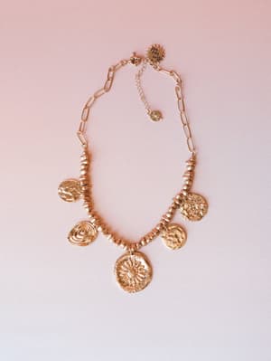 A gold coin charm necklace laid flat against a soft blush background — five textured medallion pendants hanging from a beaded and paperclip chain, with a sun motif centerpiece that feels equal parts ancient artifact and everyday magic.