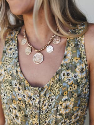 A close-up lifestyle shot of a gold coin charm necklace styled against a floral print sleeveless top — the kind of pairing that looks like it was always meant to go together.