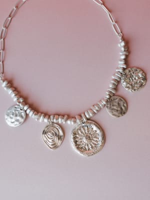 A chunky silver statement necklace laid flat on a dusty rose background — mixed beads, a paperclip chain, and five mismatched hammered coin and medallion charms, each with its own texture: a sunflower, a spiral, a cracked-earth surface, and worn cele