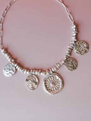 A chunky silver statement necklace laid flat on a dusty rose background — mixed beads, a paperclip chain, and five mismatched hammered coin and medallion charms, each with its own texture: a sunflower, a spiral, a cracked-earth surface, and worn cele