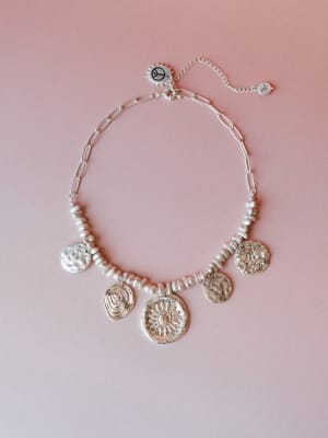 A silver coin charm necklace laid flat on a soft blush background — the kind of piece that looks like it was collected slowly, one meaningful stop at a time.