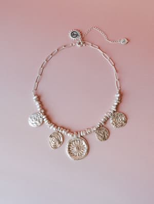 A silver coin charm necklace laid flat on a soft blush background — the kind of piece that looks like it was collected slowly, one meaningful stop at a time.