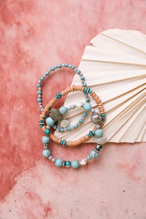 The San Antonio Bracelet Set - Turquoise, featuring boho layered bracelets with turquoise, wooden, and silver beads, is displayed in a circle on a pink marbled surface beside a folded ivory paper fan.
