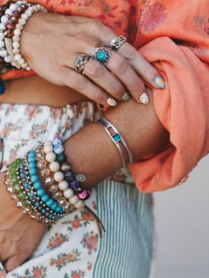 A close-up of stacked beaded bracelets and silver rings layered across both wrists, styled against a floral top and coral scarf — the kind of arm party that looks collected over time, not purchased all at once.