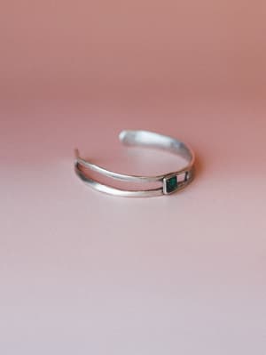A silver open-cuff bracelet with a double-band design and a small square emerald stone at center, shot on a soft blush background with gentle directional light.