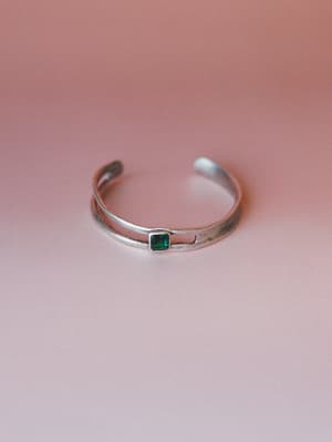 A double-band silver cuff bracelet with a small bezel-set emerald green square stone, shot on a soft dusty rose gradient background — simple and a little sentimental.