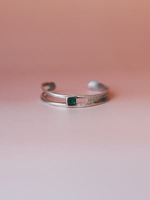 A silver cuff bracelet with a double-band open design and a single bezel-set square emerald stone, shot against a soft dusty rose gradient background — simple, collected, and quietly striking.