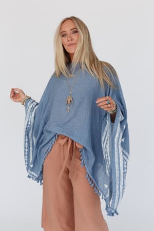 Woman with long blonde hair wears the Fallon Embroidered Sleeve Tassel Poncho in blue, featuring geometric white embroidery and tassels, paired with tan wide-leg pants and layered necklaces, standing against a plain light background.