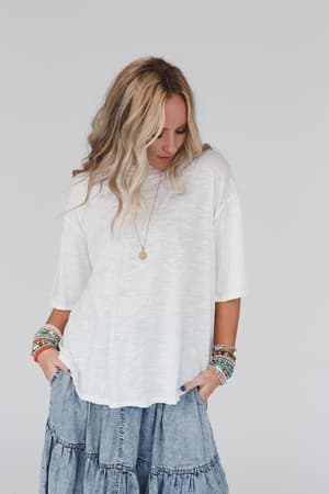 A woman with wavy blonde hair wears The Nest Your Favorite Slub Tee Shirt in Natural, paired with a tiered denim skirt, colorful beaded bracelets, and a gold pendant necklace, standing against a plain light background and looking down.