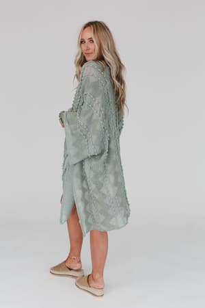 A woman with long blonde hair models the Bali Embroidered Kimono in sage, paired with tan espadrille flats and light bracelets. She stands against a plain white background, looking over her shoulder.