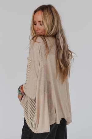 A woman with long, wavy blonde hair stands sideways, looking down. She wears the City Drifter Oversized Contrast Top in taupe with dark pants and beaded bracelets for a relaxed modern boho look against a plain light background.
