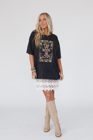 A woman with long blonde hair wears the Share The Love Graphic Tee in charcoal, paired with a white lace skirt, tan knee-high boots, silver bracelets, and a necklace, standing against a plain white background.