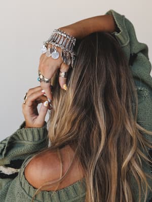 A close-up detail shot of stacked silver boho jewelry — a statement coin charm bracelet and layered rings — worn against a sage green knit sweater, with the model's long balayage hair and painted nails adding warmth and personality to the frame.