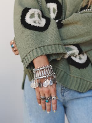 A close-up of stacked silver coin charm bracelets worn over a sage green peace sign knit sweater, layered with turquoise and silver rings — the kind of wrist stack that looks like it was collected, not curated.