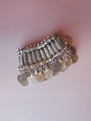 A wide silver coin bracelet laid flat on a dusty rose surface — intricate etched panels connected by rings, with rows of dangling engraved coin charms that feel like something found at a market in another country.