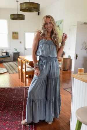 A woman with long blonde hair smiles in a bright, cozy living and dining room with wooden floors and a red rug, wearing the Harlow Wide Leg Chambray Jumpsuit - Blue and bracelets.