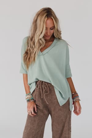 A woman with long, wavy blonde hair wears The Nest On The Go V Neck Ribbed Essential Tee in sage from Three Bird Nest and brown drawstring pants. She looks down, displaying colorful beaded bracelets on both wrists against a plain background.
