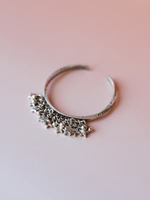 A silver cuff bracelet with dangling star charms and small ball accents rests against a soft pink background — the kind of piece that looks like it belongs on a wrist that also wears vintage rings and never takes it off.