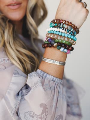 A wrist stacked with layered beaded bracelets in earthy and jewel tones alongside a silver feather cuff, raised against a soft neutral backdrop — the kind of arm party that looks collected over time, not assembled in an afternoon.