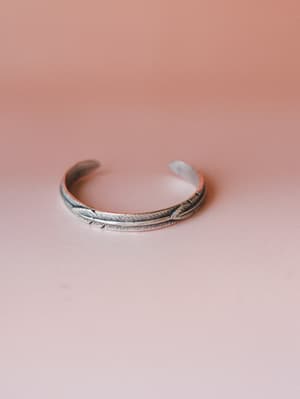 A silver cuff bracelet with an engraved feather motif rests against a soft blush-to-dusty rose gradient background — simple, earthy, and quietly personal.