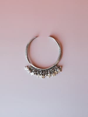A silver open-cuff bracelet with etched detailing along the band and a fringe of dangling star charms and small beads, laid flat against a soft blush pink background.