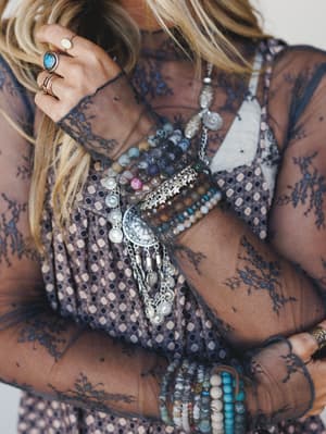 A close-up of layered boho jewelry styled on a model wearing a sheer navy lace top — stacked bead bracelets, a silver coin chain bracelet, and stone rings create that perfectly collected, not-overthought wrist stack.