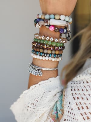 A close-up of a wrist stacked high with layered beaded and charm bracelets — stone, crystal, turquoise, and silver — worn over a cream crochet sleeve, giving off that perfectly collected, never-too-precious boho energy.