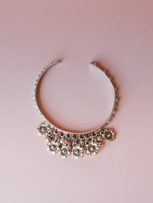 A silver open-cuff bracelet with dotted band detailing and a cluster of dangling flower charms, photographed flat against a soft dusty rose background — the kind of piece that makes a simple outfit feel intentional.