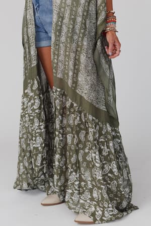 A person wearing blue denim shorts, tan pointed-toe shoes, multiple colorful bracelets on one wrist, and the Paisley Tapestry Free Flow Duster Kimono in Olive.