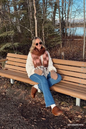 Woman sitting on a bench in a forested area wearing sunglasses and a plaid scarf.