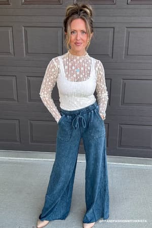 Woman wearing a white blouse with sheer details and blue jeans standing in front of a garage door.
