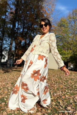 Woman in a floral dress and lace cardigan standing in a park with trees and blue sky.