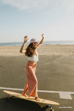 A woman in the Game On Cargo Wide Leg Pant in burnt orange, a white top, and a bucket hat balances on a longboard by the beach. The sunny scene includes the ocean and sandy shore in the background.