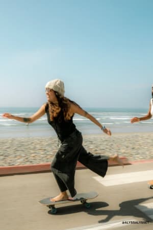 A woman wearing the Game On Cargo Wide Leg Pant in black, along with a beanie and sunglasses, skateboards barefoot on a sunny beachside path with the ocean and blue sky behind her.