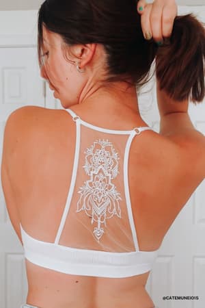 A woman with short brown hair lifts her hair, revealing the back of a white Tattoo Bralette with intricate lace and floral embroidery. She stands before white closet doors.