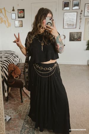 A woman with wavy hair snaps a mirror selfie, flashing a peace sign. She pairs a black crop top with The Perfect Pocketed Maxi Skirt - Black, accentuating her colorful arm tattoo. Art and patterned rugs decorate the room.