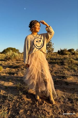 A woman stands outside on a sunny day in the Boho Ballerina Skirt - Nude, wearing a smiley face sweater, shielding her eyes with her hand amid a dry landscape and scattered shrubs.