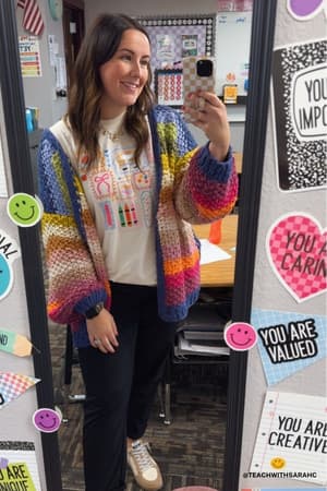 A woman takes a mirror selfie in a classroom wearing the Wrapped up in Color Knit Cardigan - Rainbow over a white T-shirt with school supply designs. The mirror features positive stickers and motivational messages.