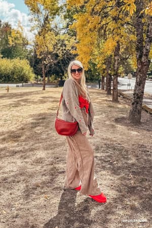 A woman with long blonde hair and sunglasses stands outdoors on dry grass, wearing So Comfy Wide Leg Pant Full Length - Mocha, a large red bow, red shoes, and carrying a red purse. Trees with yellow leaves are in the background.