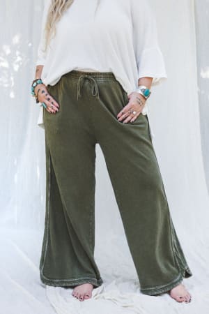 A person stands barefoot on a white fabric background, wearing the So Comfy Wide Leg Pant Cropped Length in Olive with a flowy white top. Hands with bracelets and rings are tucked into pockets, and their face is not visible.