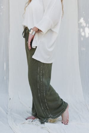 On a light fabric backdrop, a person stands barefoot in a loose white top and the So Comfy Wide Leg Pant Cropped Length in Olive, featuring a high-rise elastic waist. Their long wavy hair falls over one shoulder and they wear a silver bracelet.