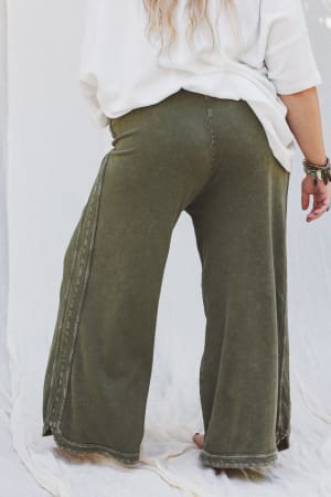 A person stands barefoot on a light fabric backdrop, wearing the So Comfy Wide Leg Pant Cropped Length in olive and a loose white top. Their long, wavy blond hair flows down their back, accessorized with bracelets on their right wrist.