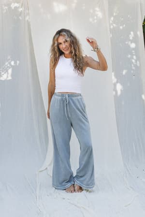 A woman with long curly hair stands barefoot on a light fabric backdrop, smiling in a white sleeveless crop top and the So Comfy Wide Leg Pant Cropped Length in Faded Blue, her right hand slightly raised for a relaxed, carefree vibe.