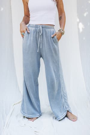 Wearing the So Comfy Wide Leg Pant Cropped Length in Faded Blue, a person stands barefoot on a light fabric background with hands in pockets, styled with a white crop top and bracelets.