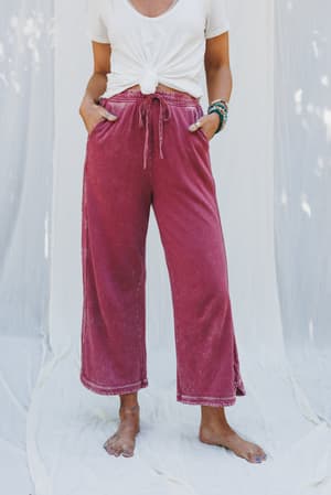A person stands barefoot on a white sheet, wearing the So Comfy Wide Leg Pant Cropped Length in Burgundy with a high-rise elastic waist and loose fit, plus a white t-shirt knotted at the front. Their hands rest casually in their pockets.